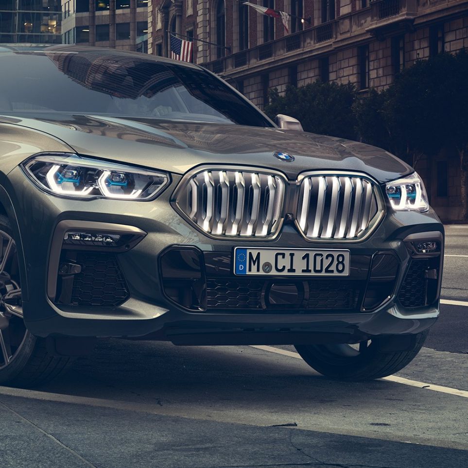 停車中の BMW X6 M50i。G06 市街地を背景に停車する BMW X6 M50i のスリークウォーター・フロントビュー。G06