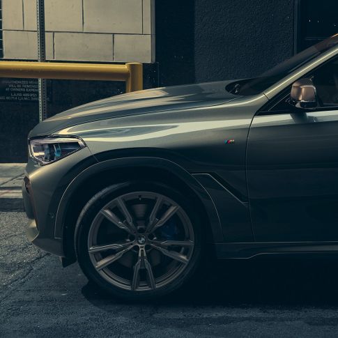 G06 市街地を背景にしたサイドビューの BMW X6 M50i に寄りかかる男性。G06