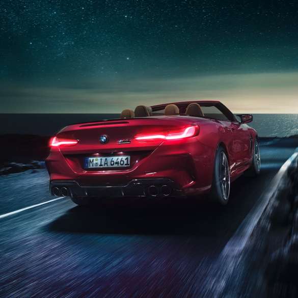 BMW M8 Competition カブリオレ、スリークウォーター・リアビュー。F91 BMW M8 Competition カブリオレのモテギ・レッド・メタリックカラー (ソフトトップを開けた状態)、夜の海岸道路を走行中のスリークウォーター・リアビュー。F91