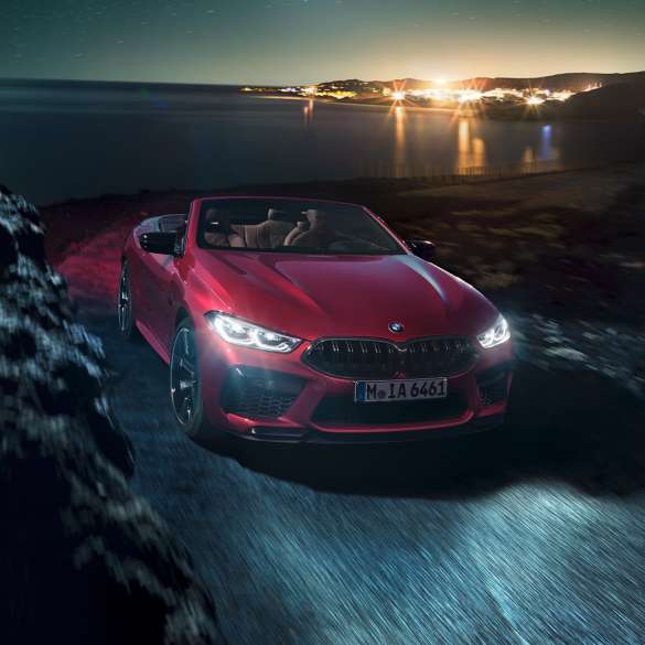 BMW M8 Competition カブリオレ、スリークォータービュー。F91 BMW M8 Competition カブリオレのモテギ・レッド・メタリックカラー (ソフトトップを開けた状態) 、夜の海岸道路上でのスリークォータービュー。F91