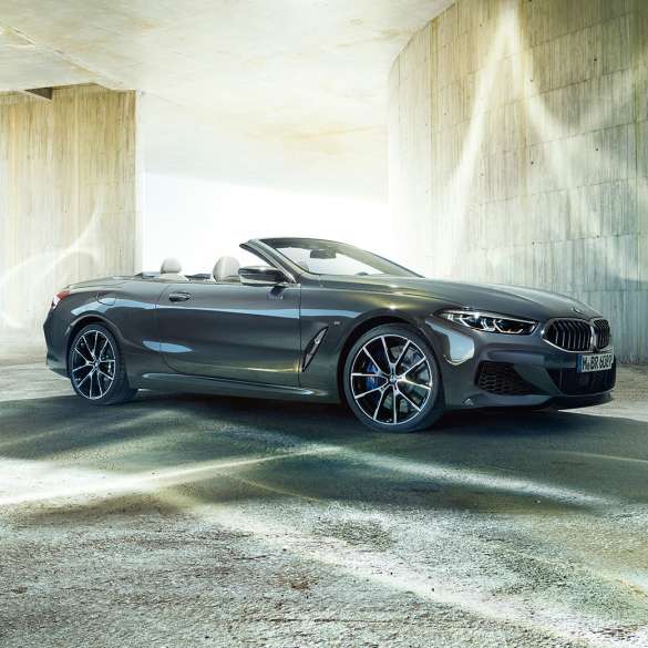 BMW M850i xDrive カブリオレ、スリークウォーター・サイドビュー。G14 BMW M850i xDrive カブリオレのドラバイト・グレー・メタリックカラー (ソフトトップを開けた状態) 、コンクリートのビルを背景にしたスリークウォーター・サイドビュー。G14