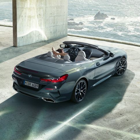 BMW M850i xDrive カブリオレ、バードアイビュー。G14 BMW M850i xDrive カブリオレのドラバイト・グレー・メタリックカラー (ソフトトップを開けた状態)、海を背に建つコンクリートのビルに駐車している場面のバードアイビュー。G14
