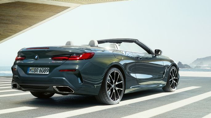 BMW M850i xDrive カブリオレ、スリークウォーター・リアビュー。G14 BMW M850i xDrive カブリオレ のドラバイト・グレー・メタリックカラー (ソフトトップを開けた状態)、海の側に駐車している状態でのスリークウォーター・リアビュー。G14