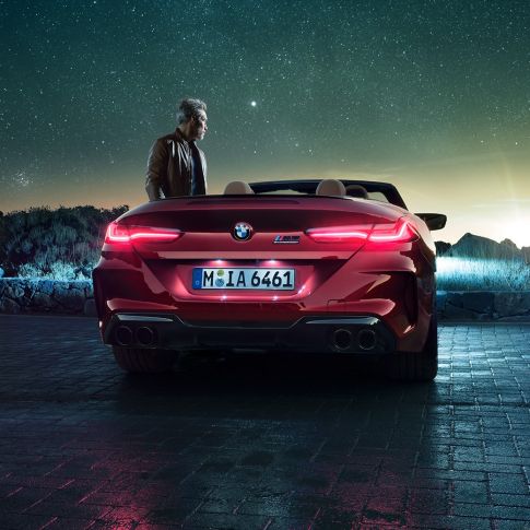 BMW M8 Competition カブリオレ、リアビュー。F91 BMW M8 Competition カブリオレのモテギ・レッド・メタリックカラー (ソフトトップを開けた状態)、星空の下での男性とのリアビュー。F91