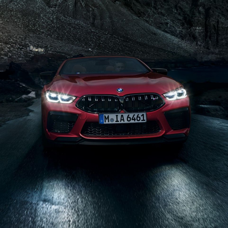 BMW M8 Competition カブリオレ、フロントビュー。F91 ヘッドライトの輝く BMW M8 Competition カブリオレのモテギ・レッド・メタリックカラー、夜間走行中のフロントビュー。F91