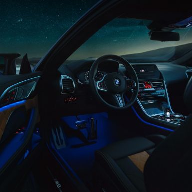 BMW M8 コンペティションクーペ、インテリア。F92 BMW M8 コンペティションクーペ、夜間に運転席側のドアを開けたビュー。Mレザーステアリングホイールとブルーの間接照明が映える。F92