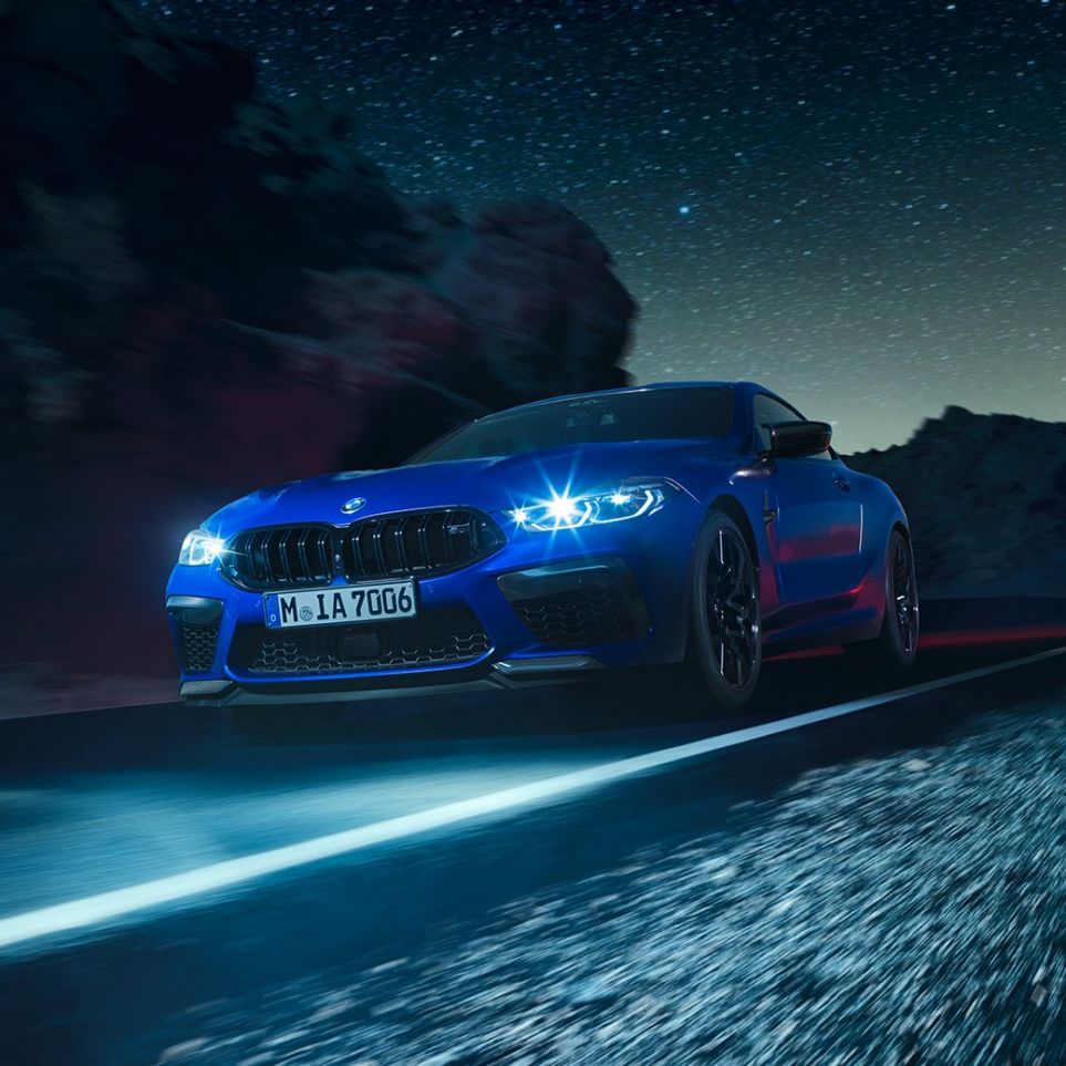 疾走するBMW M8 コンペティションクーペ、スリークォーターフロントビュー。F92 BMW M8 コンペティションクーペ(塗色マリーナベイ ブルーメタリック)。夜間走行できらめくLEDヘッドライトが印象的なスリークォーターフロントビュー。F92