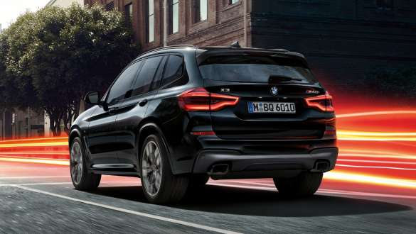 BMW X3 M40i、スリークウォーター・リアビュー。 BMW X3 M40i のブラック・サファイア・メタリックカラー、走行中、エクステリア、市街を背景にライトを点灯させているスリークウォーター・リアビュー。