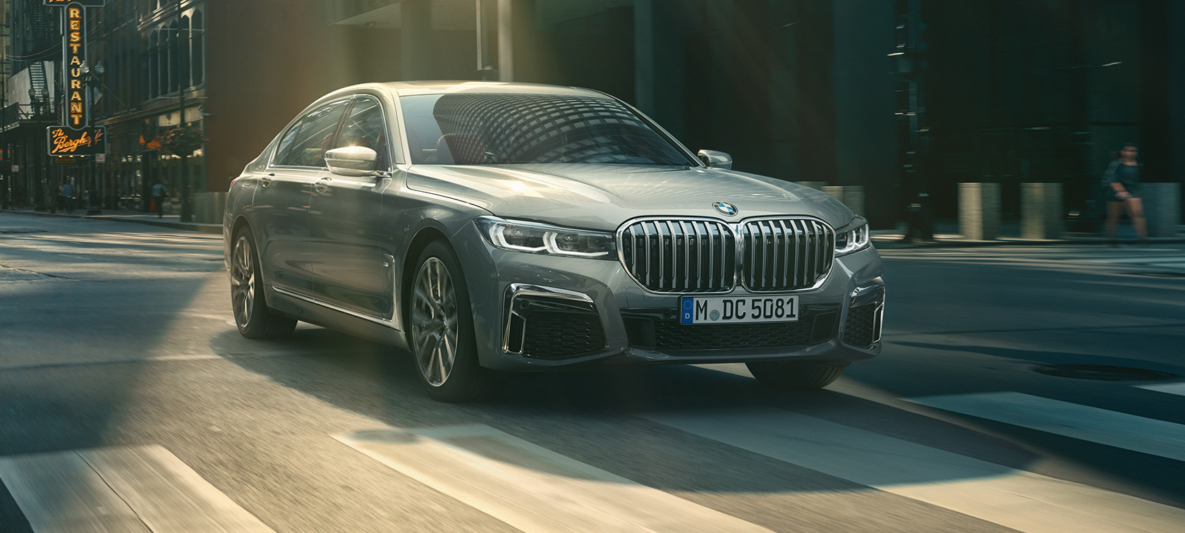 走行中のグレーのBMW 7シリーズ セダン 斜めからのフロント・ビュー 都市風景の中を走行するグレーのBMW 7シリーズ セダンの斜めからのフロント・ビュー