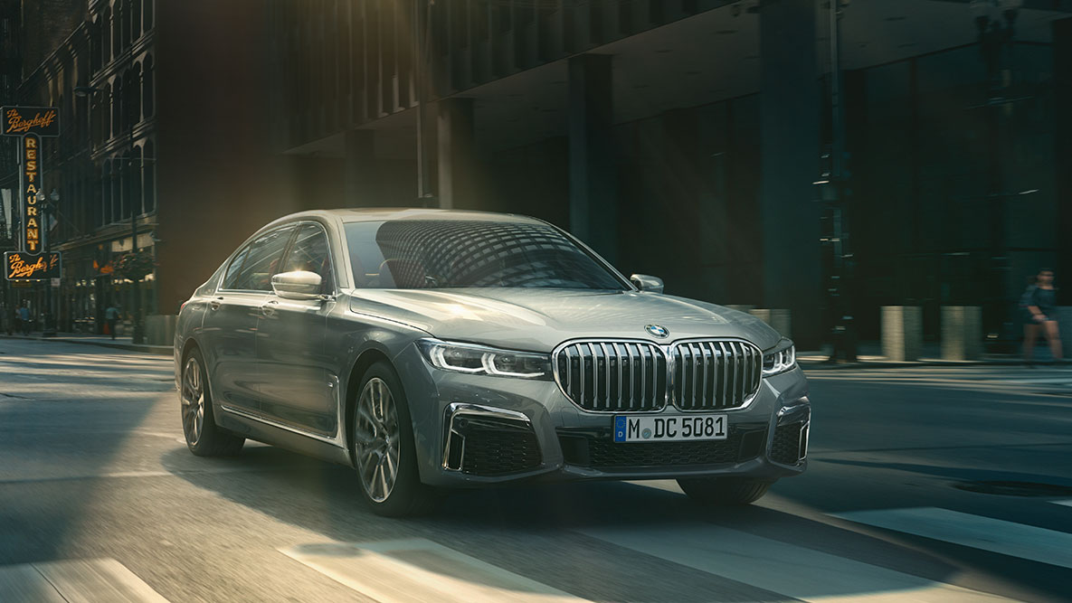 ドライビング・アシスト・プロフェッショナル:都市風景を背に走行するBMW 7シリーズ セダンの斜めからのフロント・ビュー BMW 7シリーズ セダン ドライビング・アシスト・プロフェッショナル