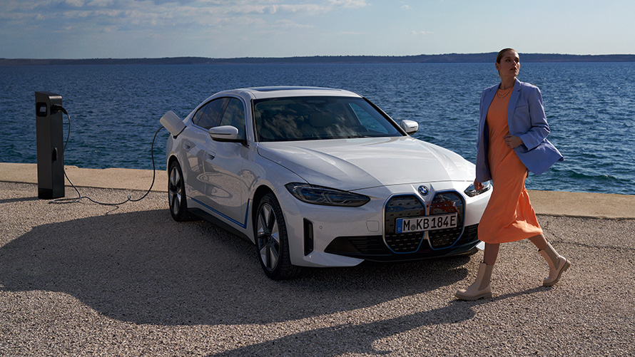 斜めからのフロント・ビュー、BMW i4 eDrive40 G26 2021、充電ステーションにて BMW i4 eDrive40 G26 2021 ミネラル・ホワイトの前にいる女性、斜めからのフロント・ビュー、湖の辺りの充電ステーションにて