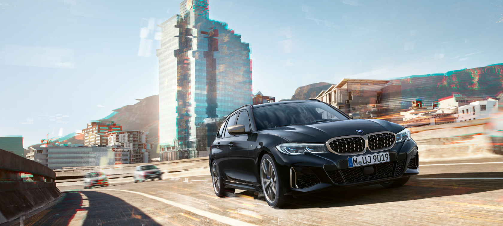 都市を背景にドライビング中の斜め前からの姿 BMW M340i xDrive ツーリング、都市を背景にドライビング中の斜め前からの姿