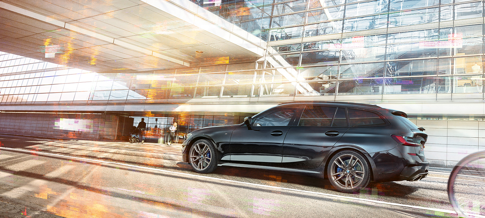 BMW M340i xDrive ツーリング、走行中の側面 BMW M340i xDrive ツーリング、都会のガラス張りビル前を走行中の側面