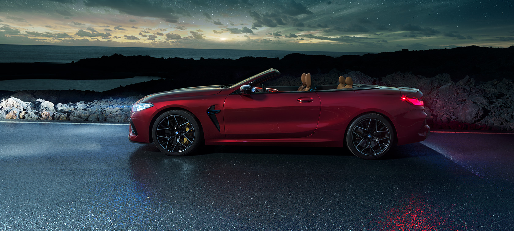 BMW M8 Competition カブリオレ、サイドビュー。 BMW M8 Competition カブリオレのモテギ・レッド・メタリックカラー (ソフトトップを開けた状態)、星空の下の海岸道路上でのサイドビュー。