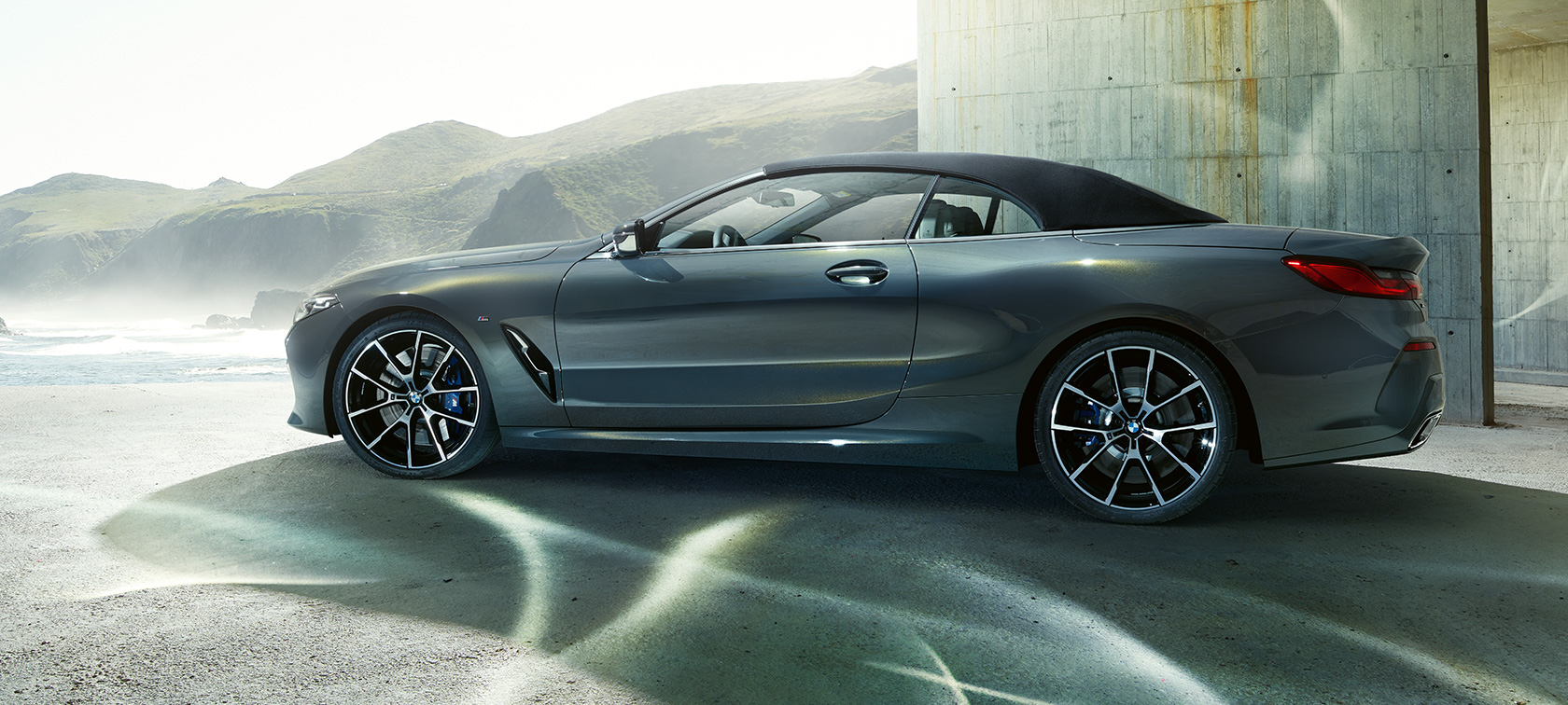 BMW M850i xDrive カブリオレ、サイドビュー。 BMW M850i xDrive カブリオレのドラバイト・グレー・メタリックカラー (ソフトトップを閉めた状態)、丘を背景にしたサイドビュー。