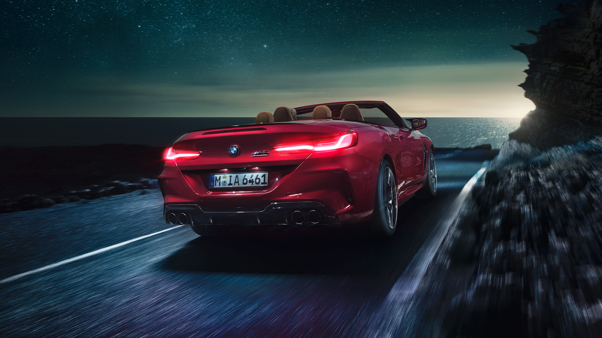 BMW M8 Competition カブリオレ、スリークウォーター・リアビュー。F91 BMW M8 Competition カブリオレのモテギ・レッド・メタリックカラー (ソフトトップを開けた状態)、夜の海岸道路を走行中のスリークウォーター・リアビュー。F91