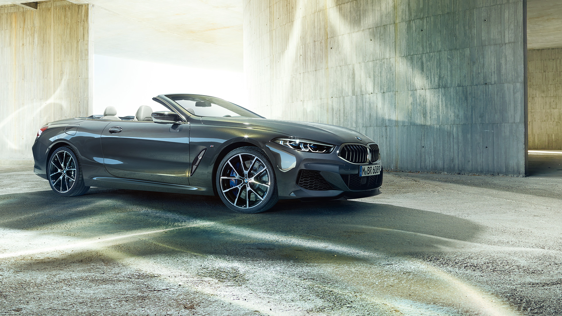 BMW M850i xDrive カブリオレ、スリークウォーター・サイドビュー。G14 BMW M850i xDrive カブリオレのドラバイト・グレー・メタリックカラー (ソフトトップを開けた状態) 、コンクリートのビルを背景にしたスリークウォーター・サイドビュー。G14