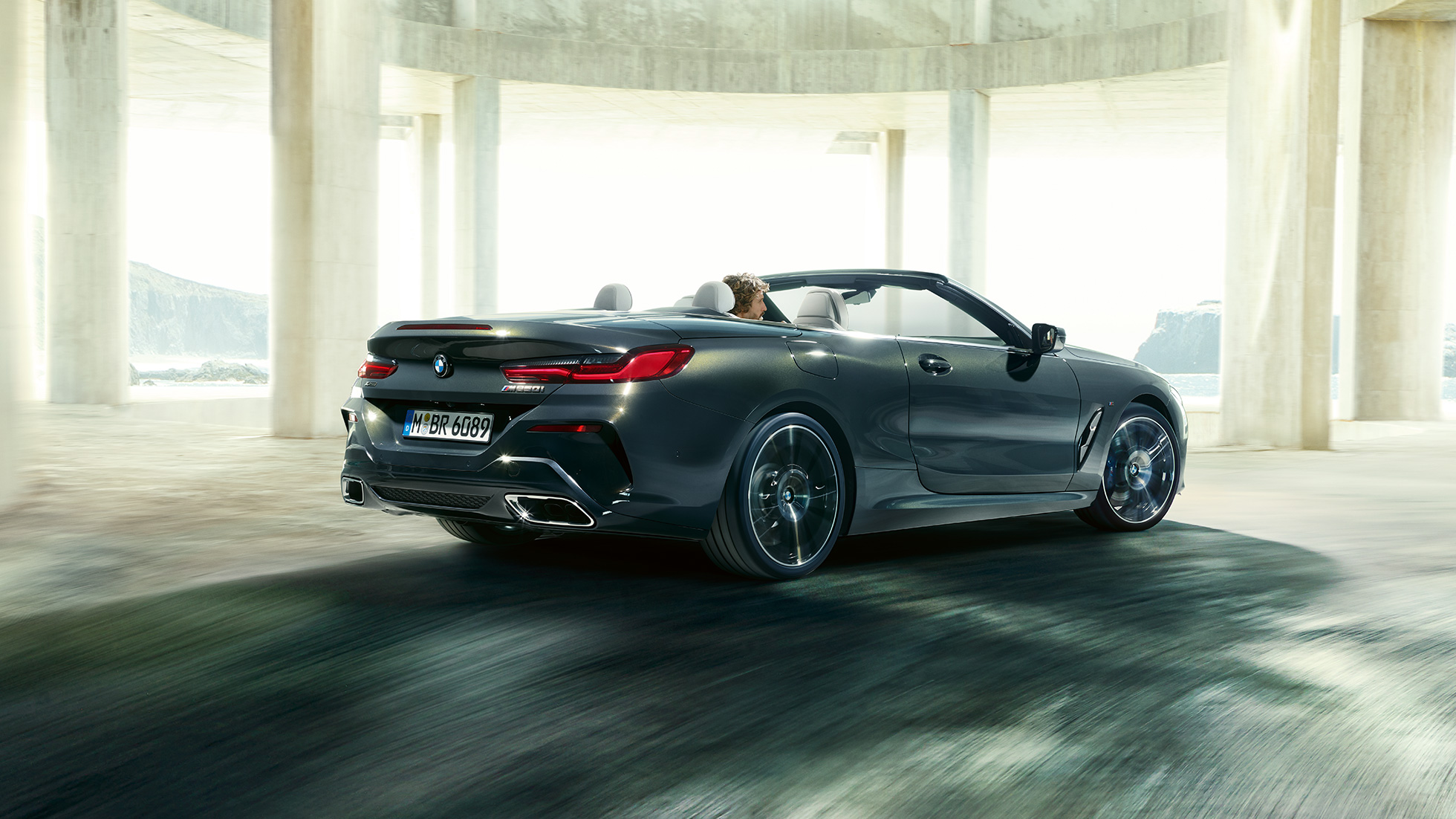 BMW M850i xDrive カブリオレ、スリークウォーター・リアビュー。G14 BMW M850i xDrive カブリオレのドラバイト・グレー・メタリックカラー (ソフトトップを開けた状態) 、コンクリートのビル内を走り抜けている時のスリークウォーター・リアビュー。G14