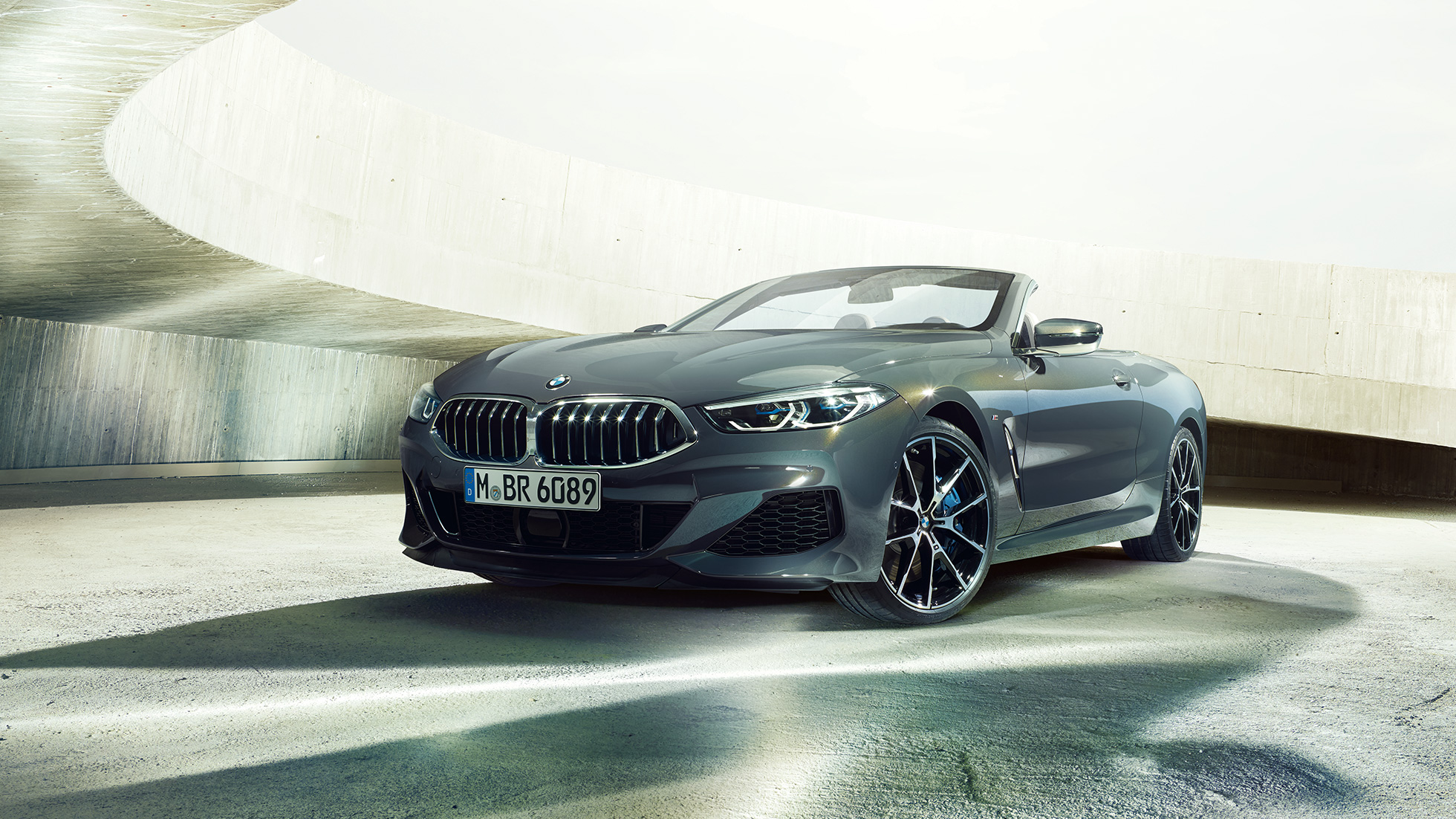 BMW M850i xDrive カブリオレ、スリークォータービュー。G14 BMW M850i xDrive カブリオレのドラバイト・グレー・メタリックカラー (ソフトトップを開けた状態)、コンクリートのビル内でのスリークォータービュー。G14