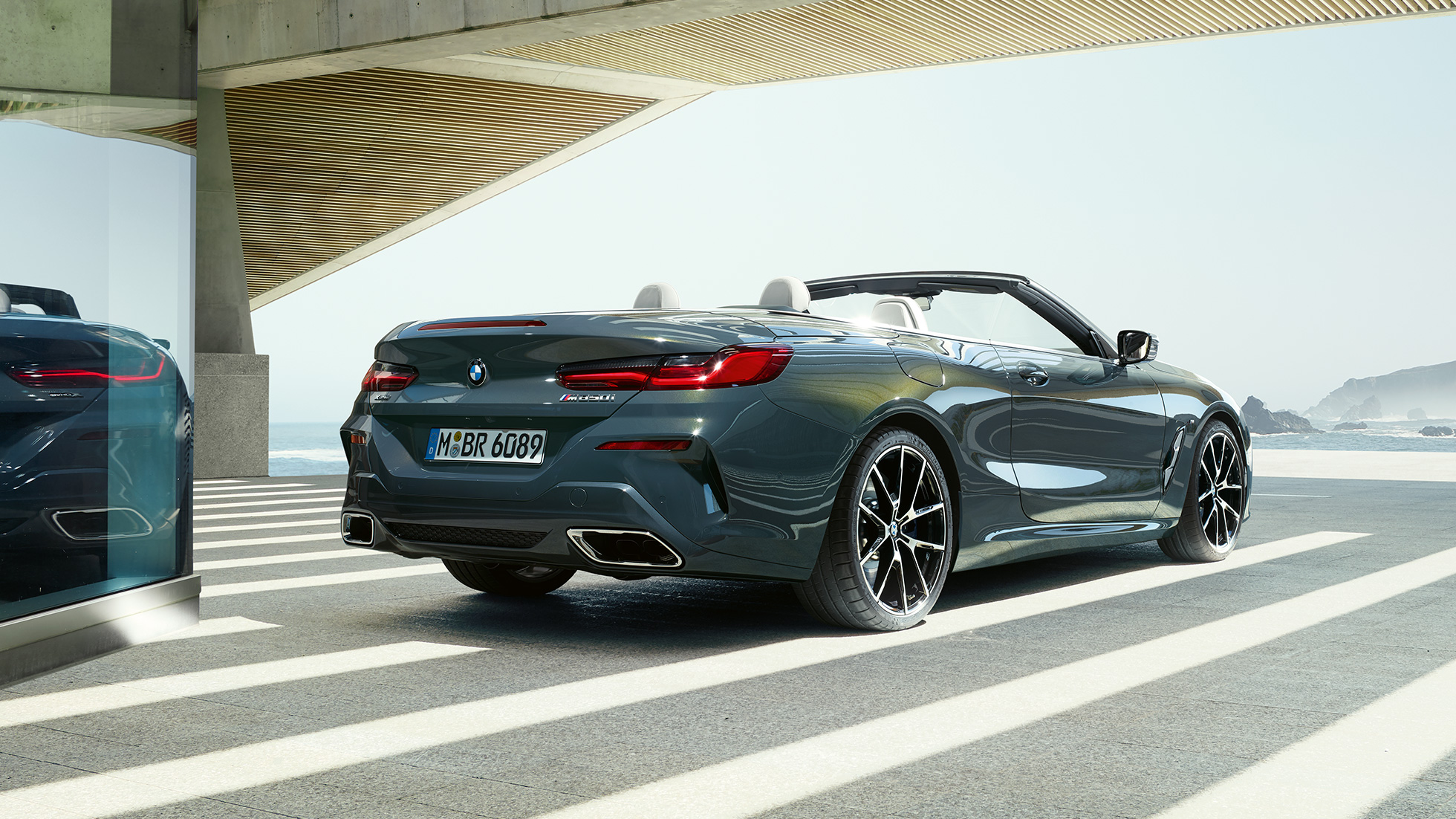 BMW M850i xDrive カブリオレ、スリークウォーター・リアビュー。G14 BMW M850i xDrive カブリオレ のドラバイト・グレー・メタリックカラー (ソフトトップを開けた状態)、海の側に駐車している状態でのスリークウォーター・リアビュー。G14