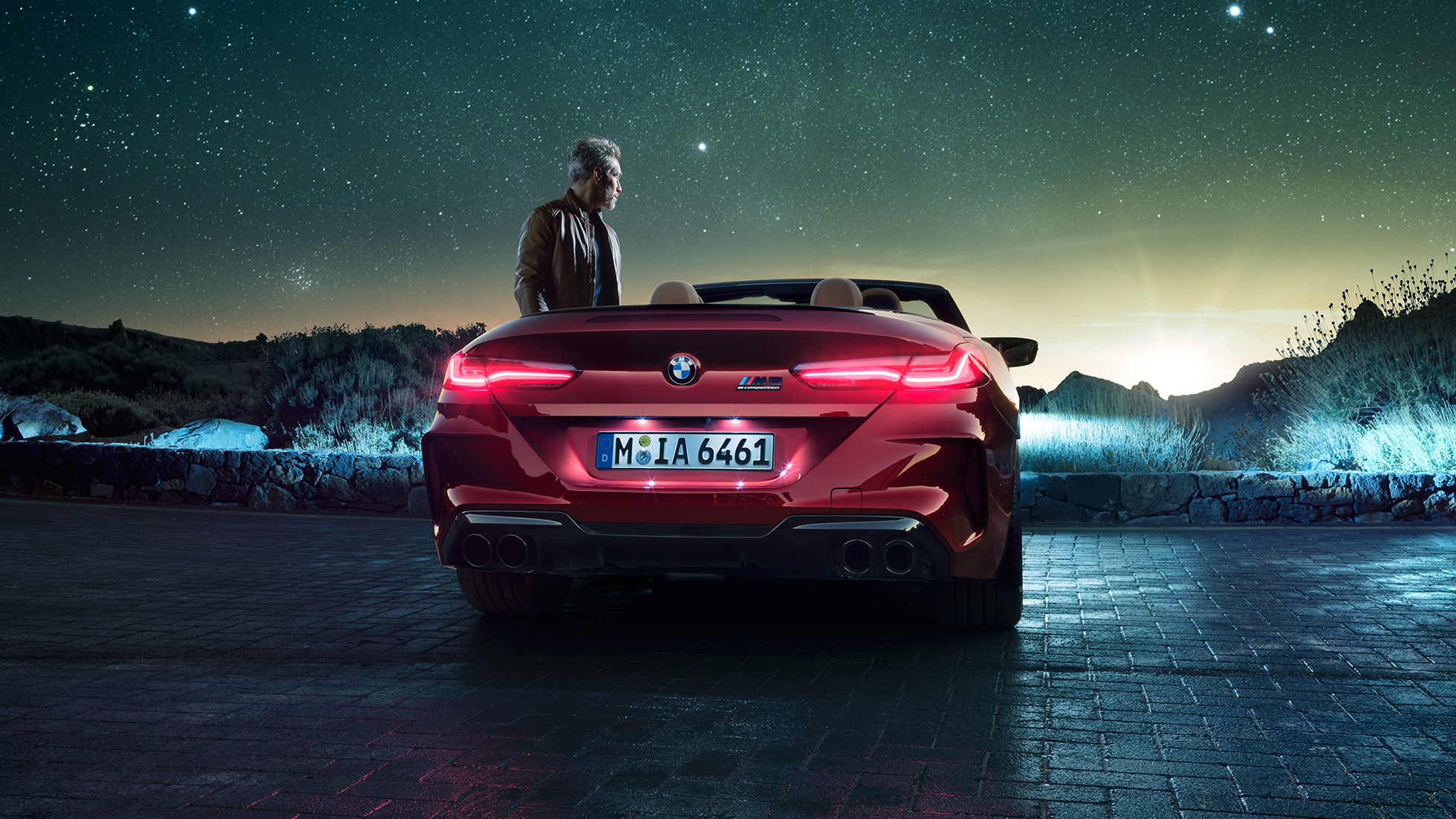 BMW M8 Competition カブリオレ、リアビュー。F91 BMW M8 Competition カブリオレのモテギ・レッド・メタリックカラー (ソフトトップを開けた状態)、星空の下での男性とのリアビュー。F91
