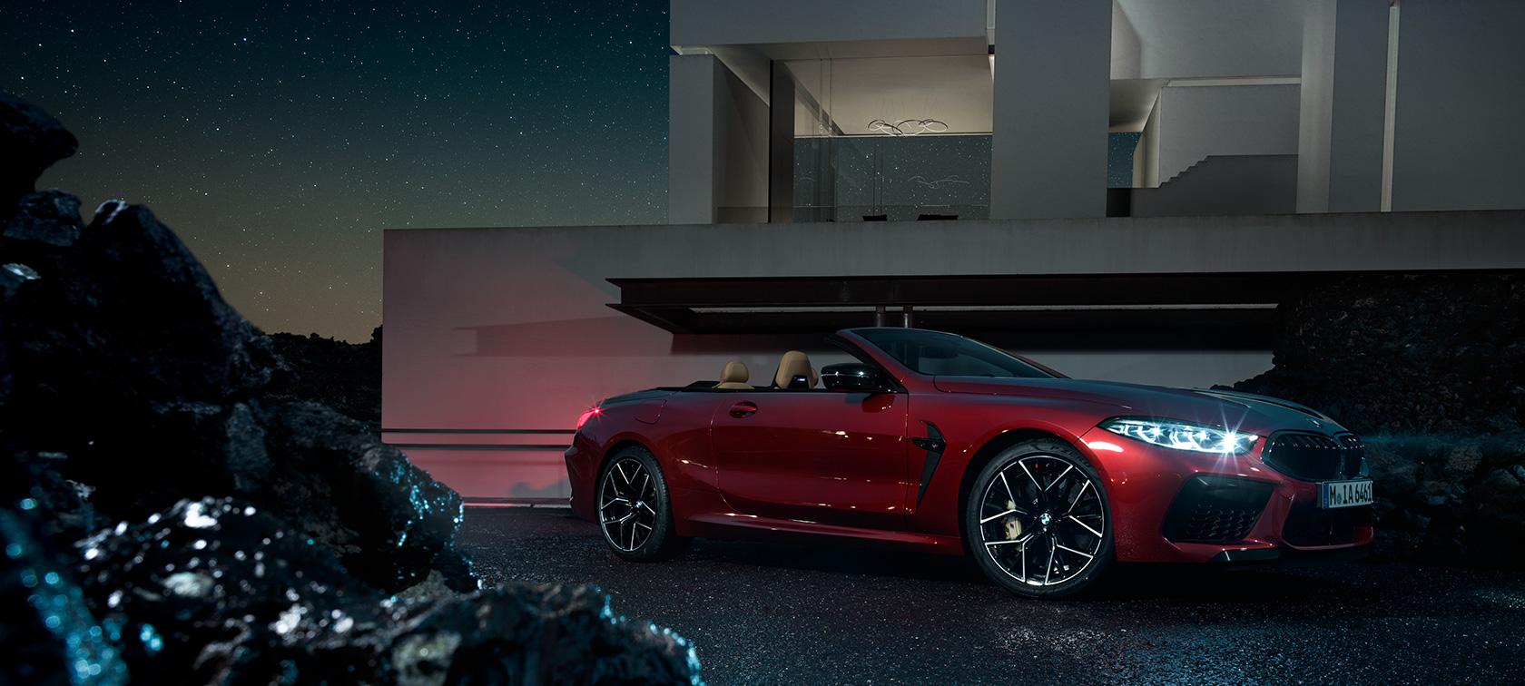 BMW M8 コンペティション カブリオレ、スリークウォーター・サイドビュー。 BMW M8 コンペティション カブリオレのモテギ・レッド・メタリックカラー、夜の別荘を背景にしたスリークウォーター・サイドビュー。