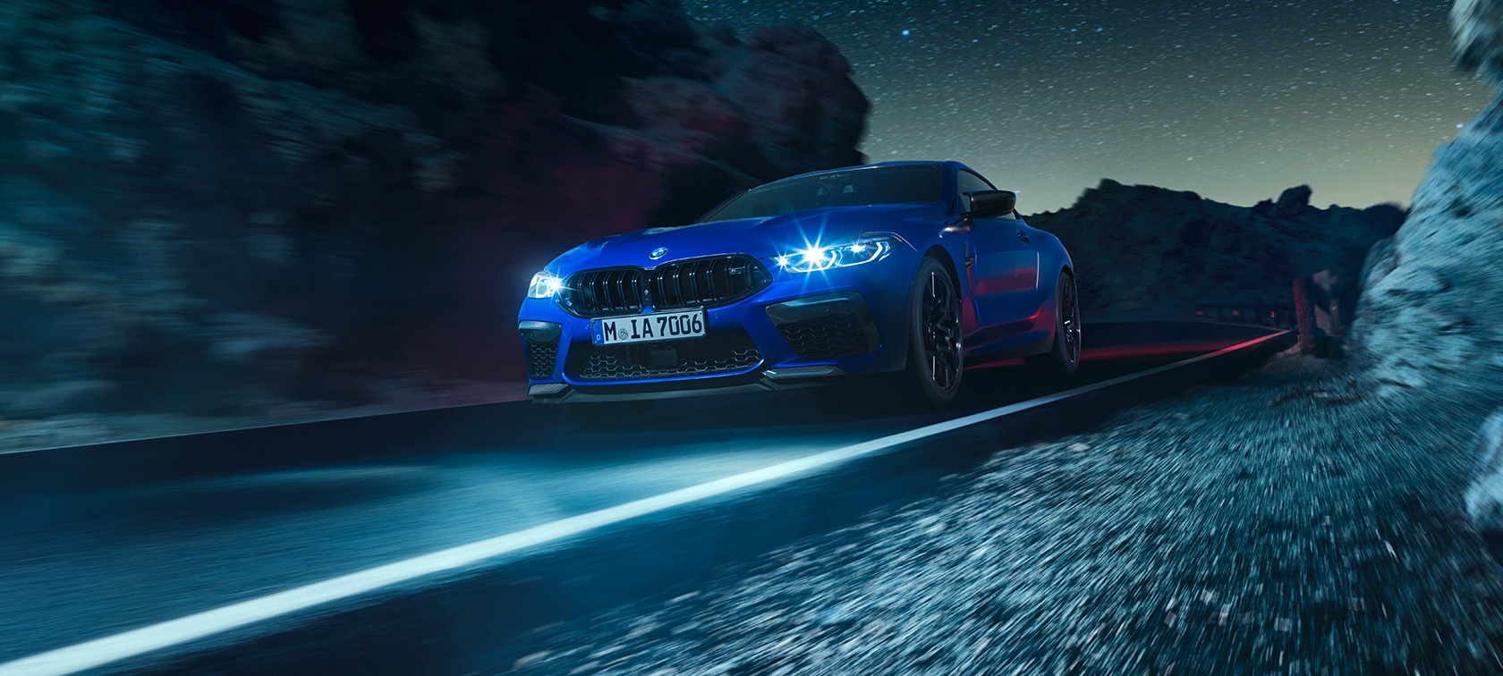 BMW M8 コンペティションクーペ、スリークォーターフロントビュー。 BMW M8 コンペティションクーペ(塗色マリーナベイ ブルーメタリック)。夜間走行のスリークォーターフロントビュー。