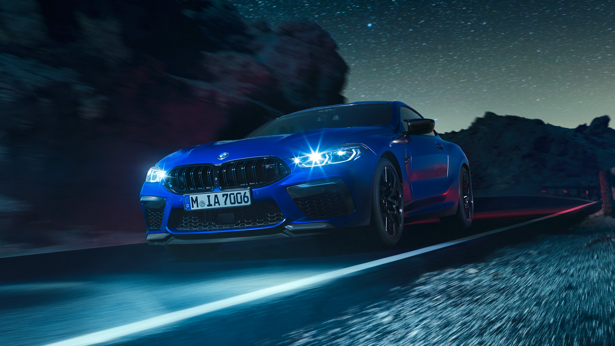 疾走するBMW M8 コンペティションクーペ、スリークォーターフロントビュー。F92 BMW M8 コンペティションクーペ(塗色マリーナベイ ブルーメタリック)。夜間走行できらめくLEDヘッドライトが印象的なスリークォーターフロントビュー。F92