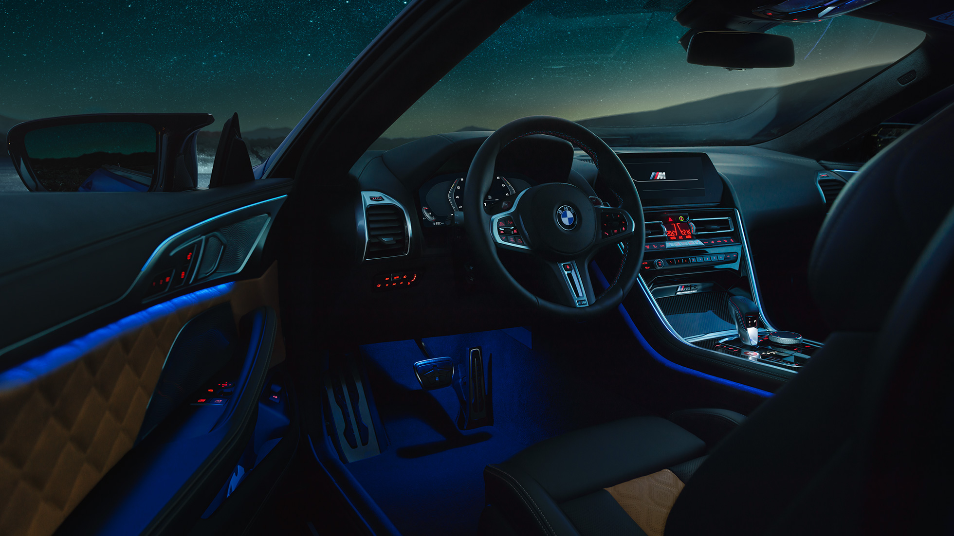 BMW M8 コンペティションクーペ、インテリア。F92 BMW M8 コンペティションクーペ、夜間に運転席側のドアを開けたビュー。Mレザーステアリングホイールとブルーの間接照明が映える。F92
