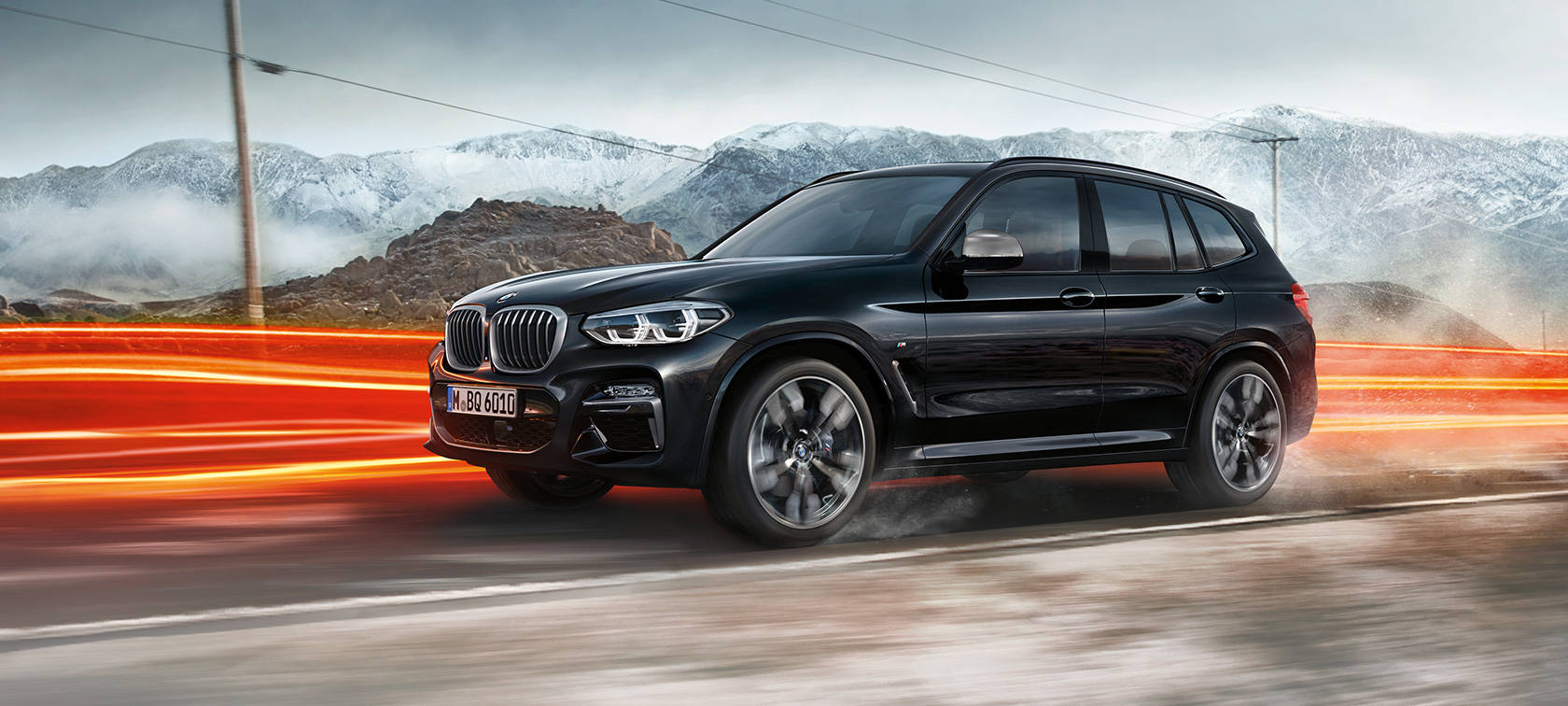 BMW X3 M40d、サイドビュー。 BMW X3 M40d のブラック・サファイア・メタリックカラー、走行中、エクステリア、山の風景をバックにライトを点灯させているサイドビュー。