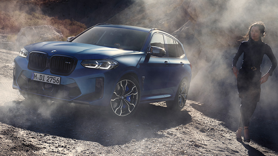 BMW X3 M Competition F97 LCI フェイスリフト 2021 フローズン・マリーナ・ベイ・ブルー・メタリック 斜めからのフロント・ビュー 女性モデルとともに石の多い地形に停車中
