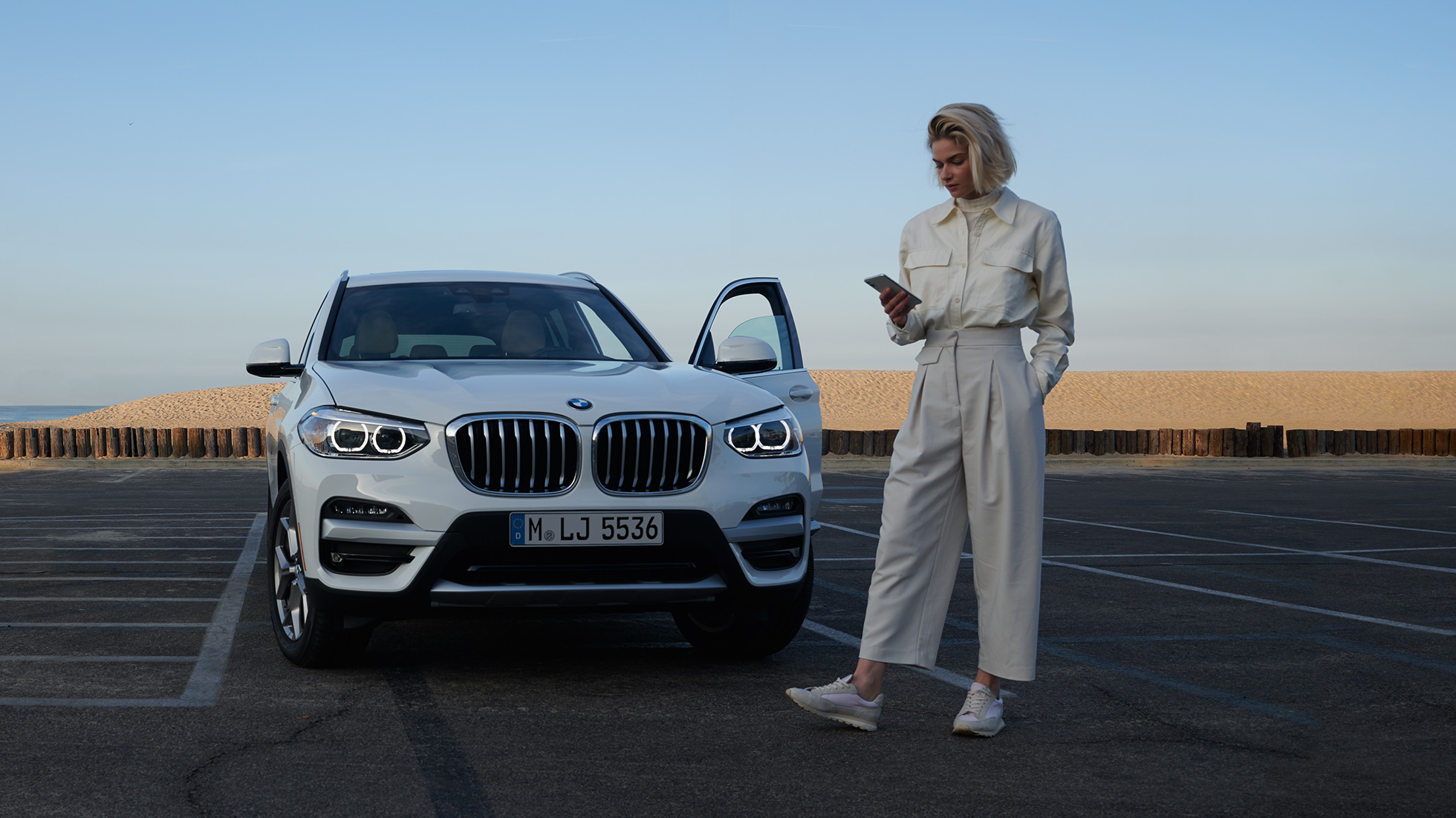 スマートフォンでMy BMW Appを操作している女性 ホワイトのBMW X5を背景にスマートフォンのBMWコネクテッド・ドライブからMy BMW Appを操作している女性